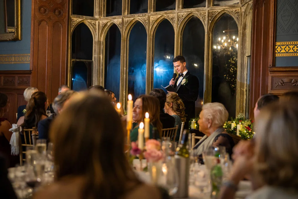 A groom in a velvet tuxedo delivers his speech during a candlelit wedding reception in a historic hall with large arched windows.