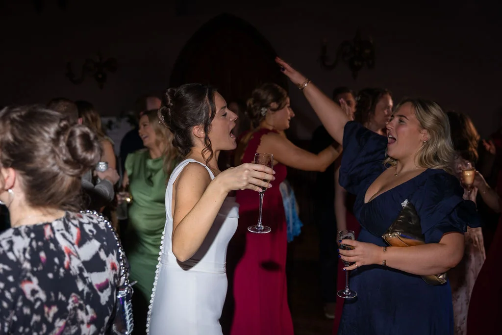 A bride in a white gown and wedding guests celebrate on a lively, crowded dance floor holding champagne glasses.