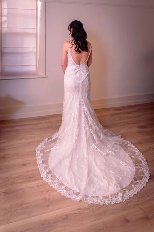 A bride stands by a bright window, showcasing the intricate lace detailing and elegant train of her wedding dress from behind.