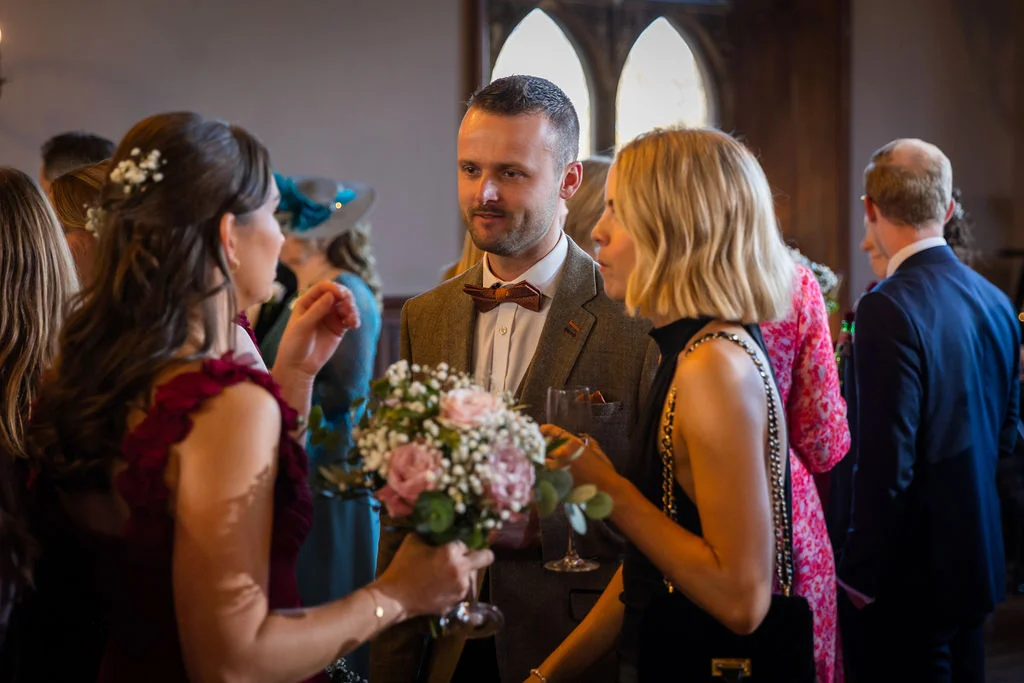 Wedding guests mingle and converse during a drinks reception inside a venue with arched windows.