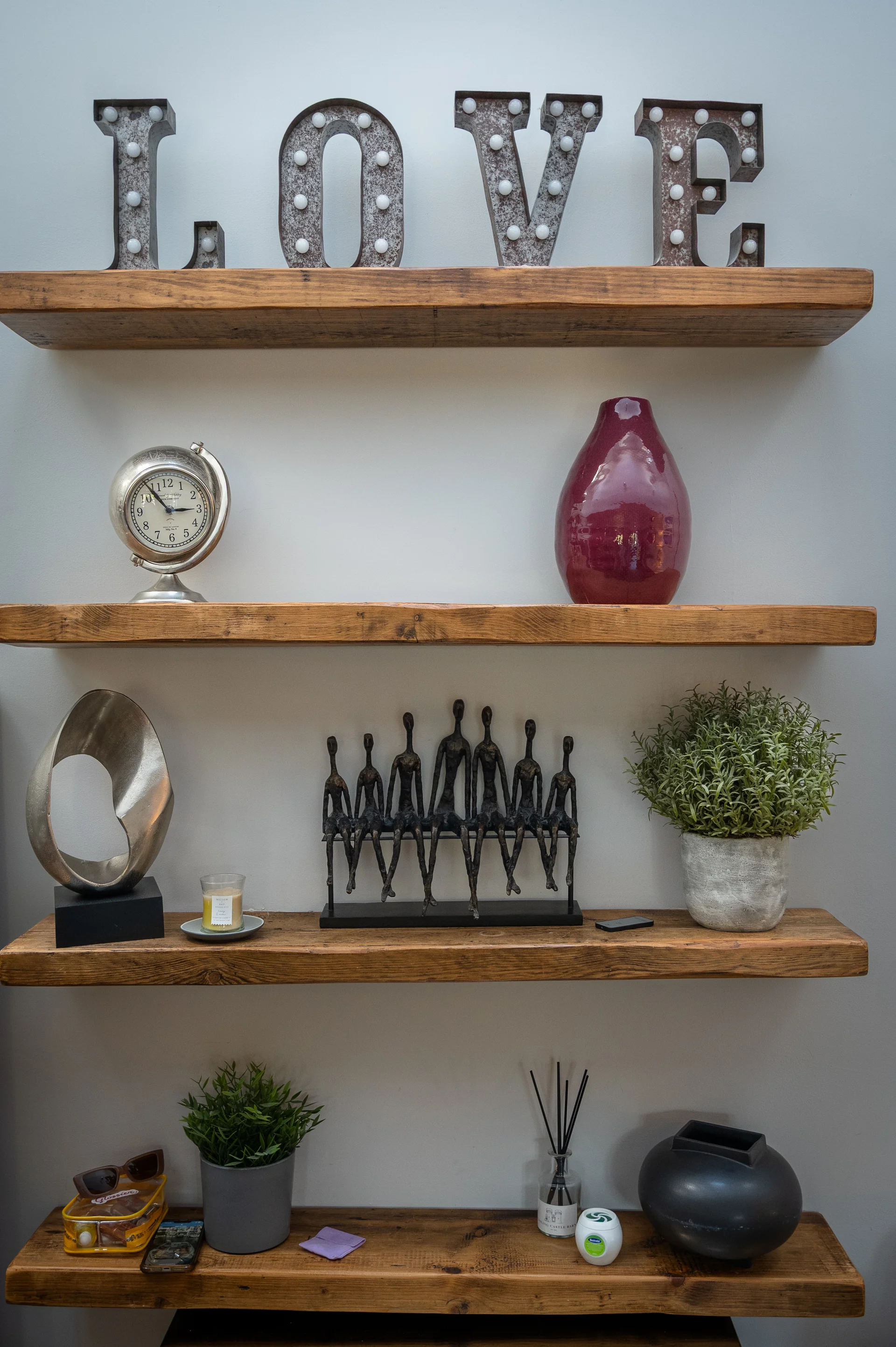 A close-up view of four rustic wooden shelves decorated with a lighted love sign, sculptures, potted plants, and various ornaments against a plain wall.