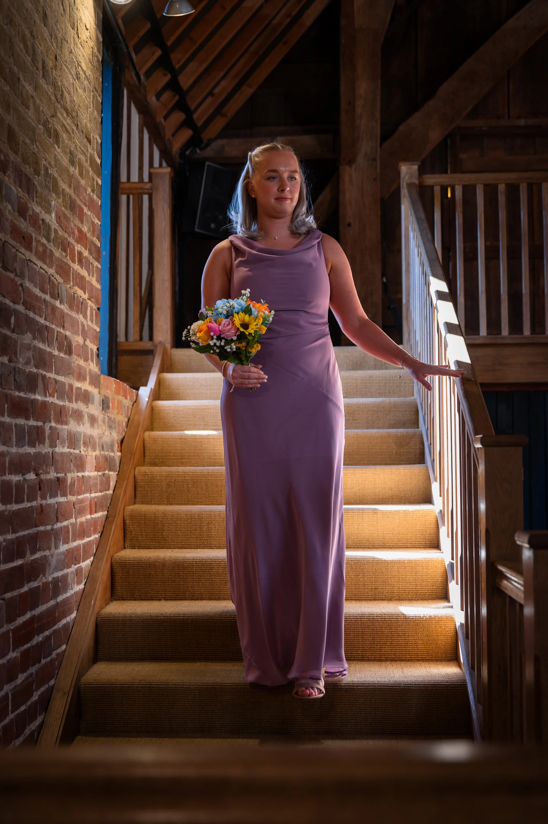 A bridesmaid in a sleek pink dress and holding a colourful bouquet descends a wooden staircase inside an exposed brick barn.