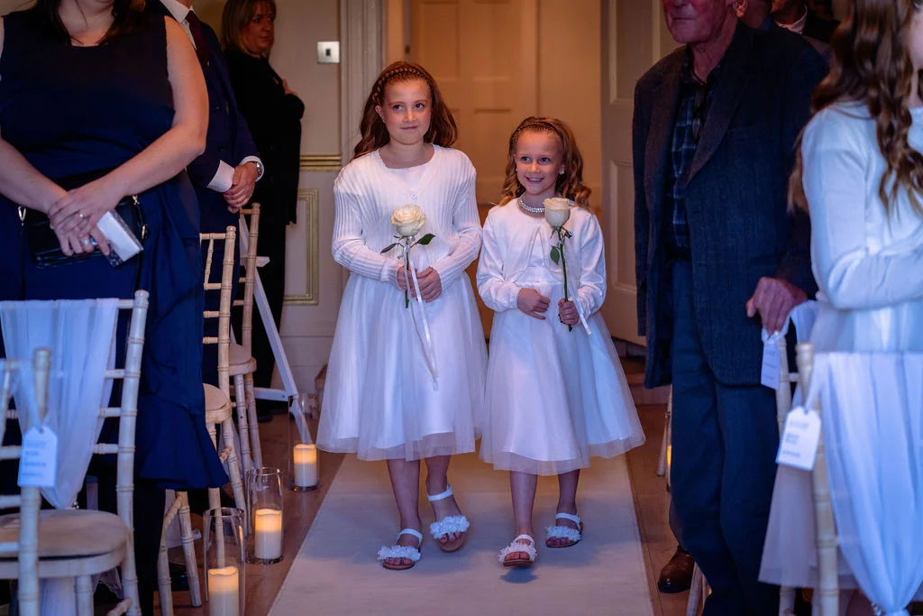 Two young flower girls in white dresses holding single white roses walk down the candlelit aisle during the wedding ceremony.