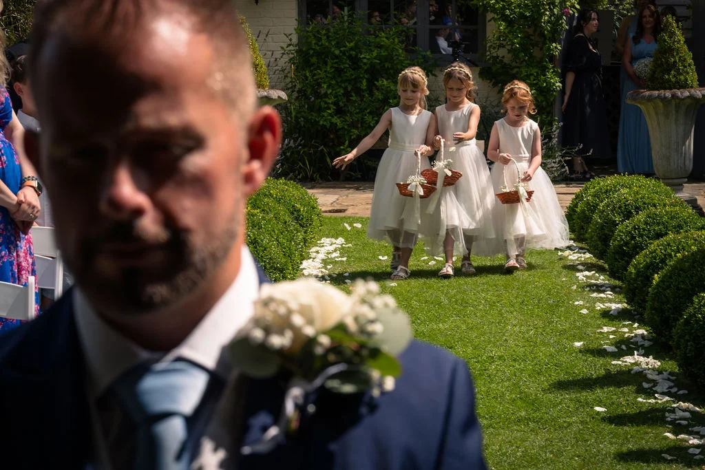 An emotional groom waits at the end of a garden aisle while three flower girls in white dresses walk towards him scattering petals.