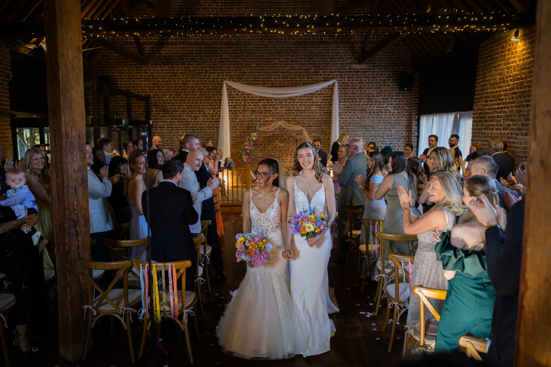 Two smiling brides walk hand in hand down the aisle of a rustic brick barn venue as cheering guests applaud them.