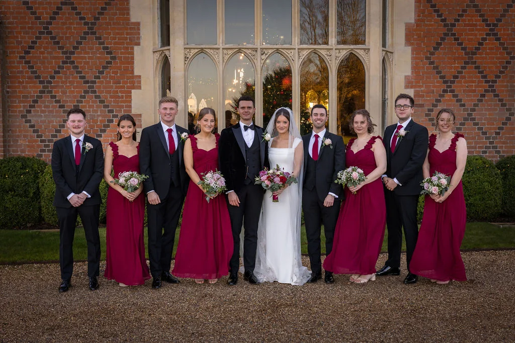 A newlywed couple poses proudly outdoors with their wedding party of four groomsmen in black tuxedos and four bridesmaids in floor-length burgundy gowns in front of a grand manor house.