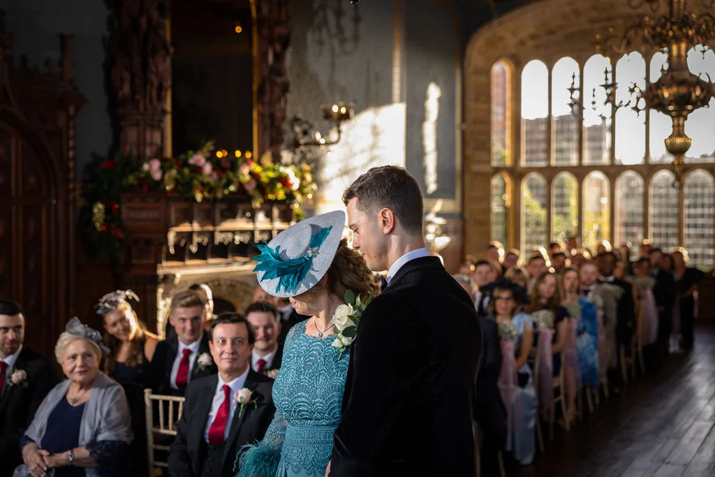 A groom and a guest wearing a feathered fascinator stand at the front of a grand, sunlit historic hall during a wedding ceremony.