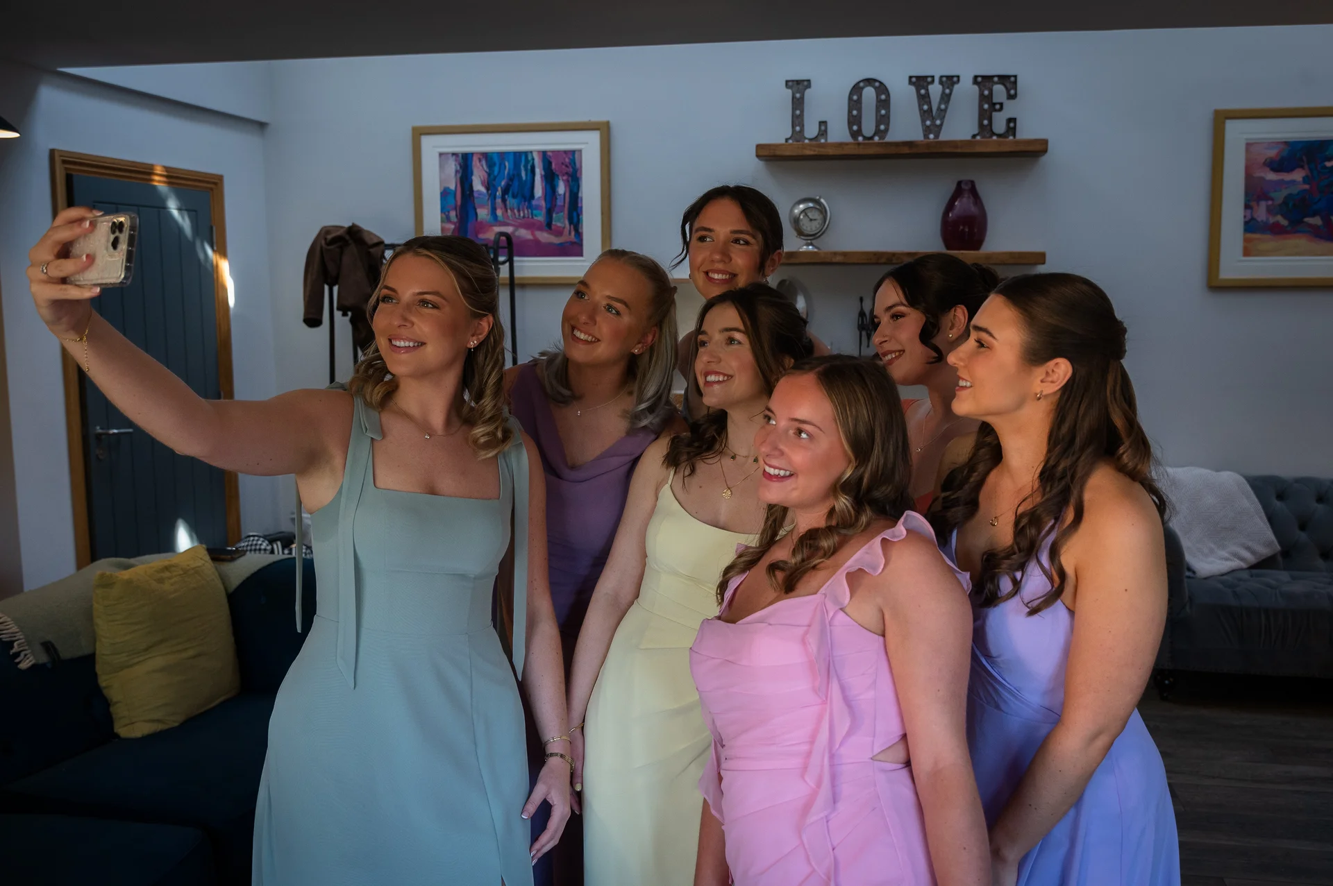 A group of smiling bridesmaids wearing pastel-coloured dresses gather indoors to take a group selfie together.