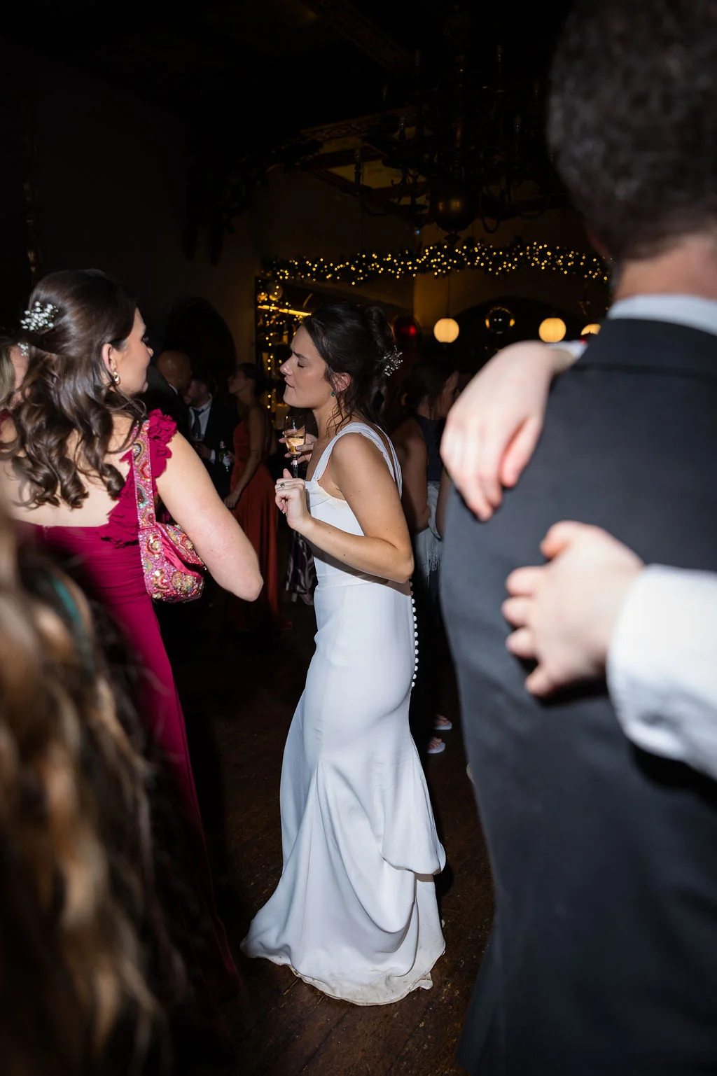 A bride in a sleek white gown dances joyfully on a crowded wooden dance floor during the evening wedding reception.