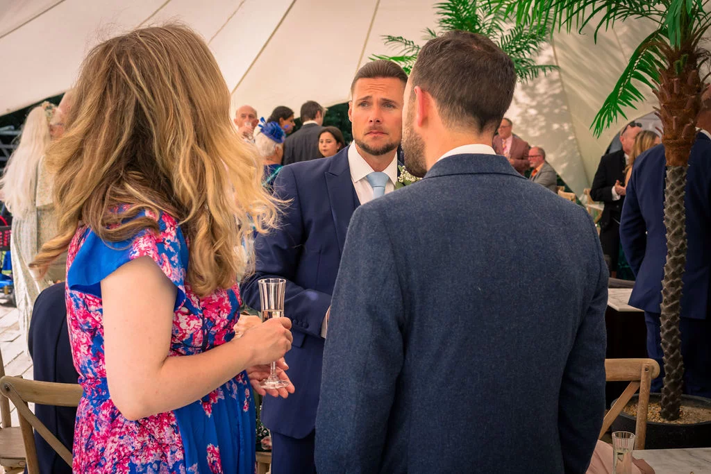 Wedding guests mingle and converse inside a bright marquee during the drinks reception, with a woman in a floral dress holding a glass of champagne in the foreground.