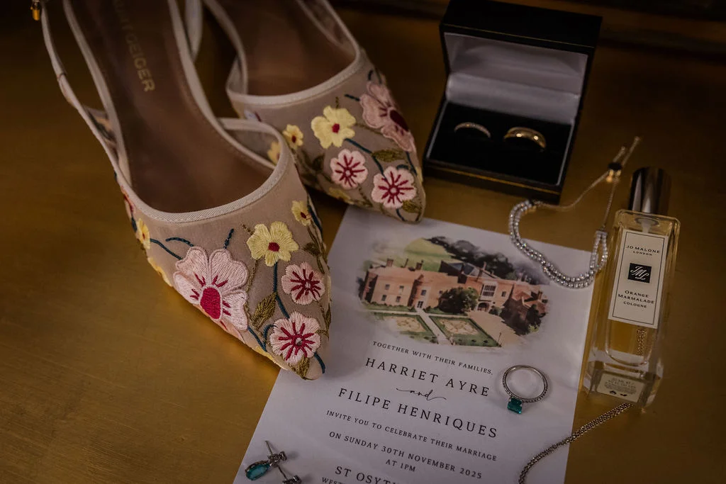 A bridal flat lay featuring embroidered floral wedding shoes, an invitation suite with a venue illustration, wedding bands in a box, an engagement ring, jewellery, and a perfume bottle.