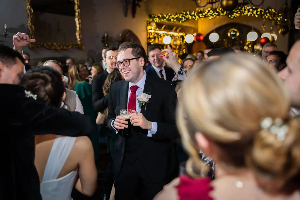 A smiling guest in a black suit and red tie holds a drink on a crowded, festive dance floor illuminated by fairy lights.