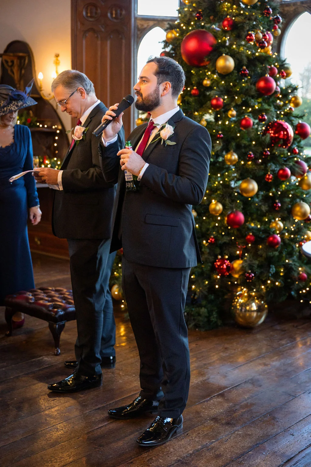 A groom delivers a speech into a microphone next to a large decorated Christmas tree while another speaker holds notes nearby during a festive winter wedding reception.