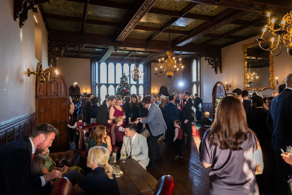 Wedding guests mingle and converse inside a historic stately home reception room featuring dark wood panelling, ornate chandeliers, and a decorated Christmas tree in the background.