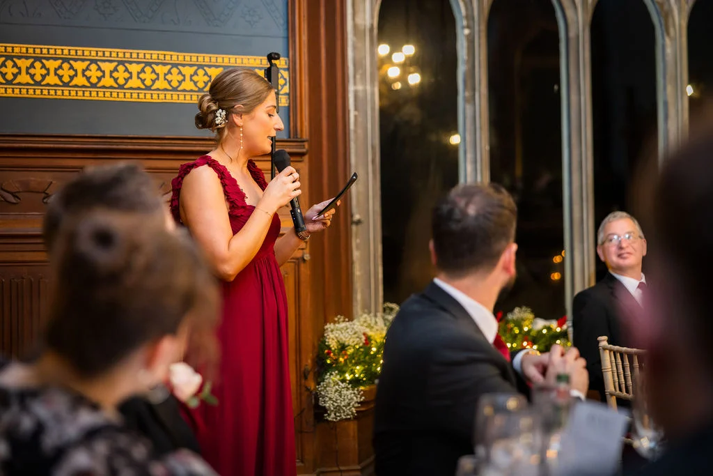 A bridesmaid in a burgundy dress delivers a speech into a microphone during the wedding reception in a wood-panelled hall.