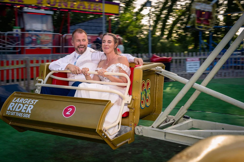 A newlywed couple laughing and riding a vintage funfair ride together during their wedding reception.