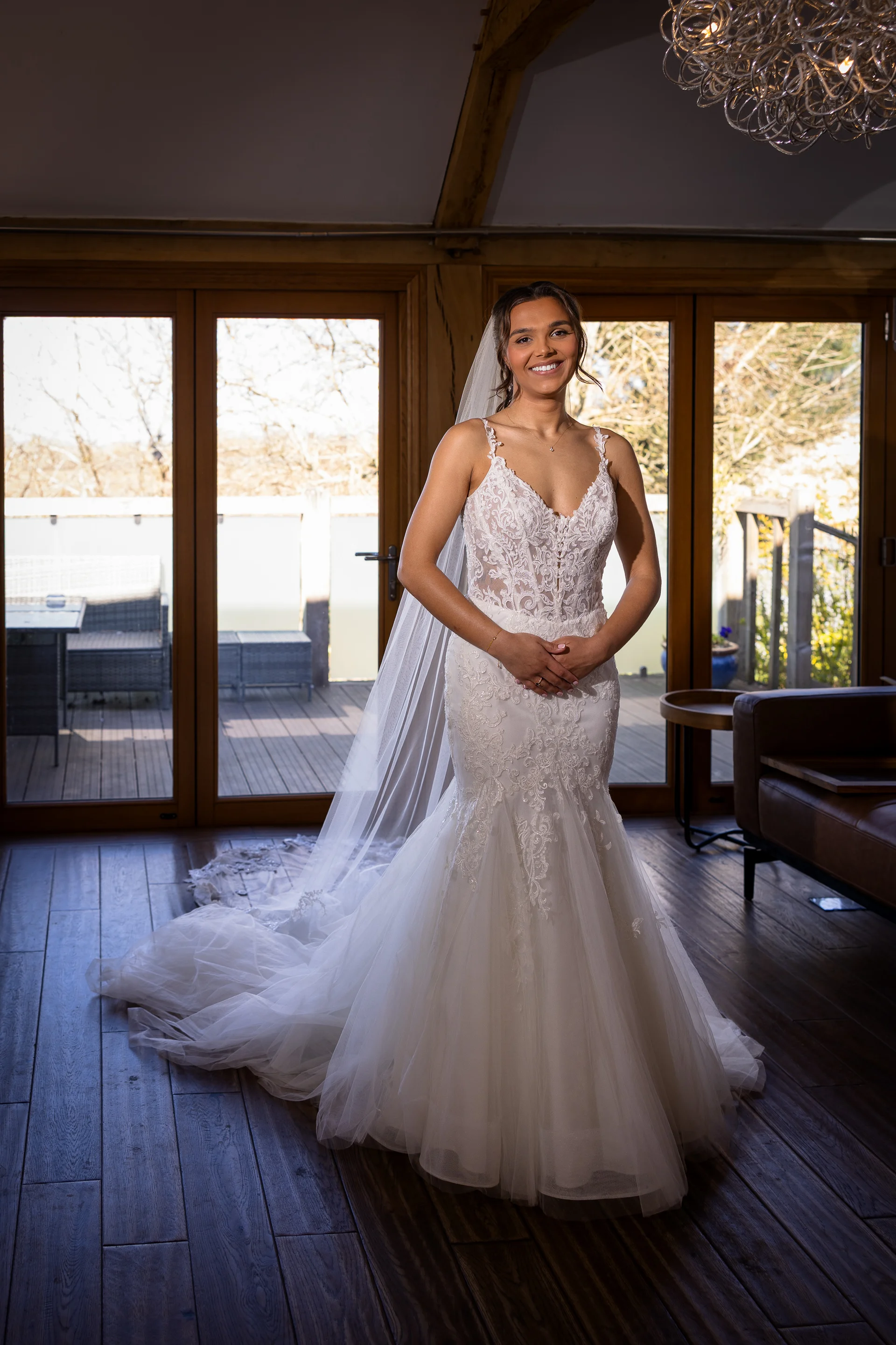 A smiling bride stands indoors wearing a lace mermaid style wedding dress with spaghetti straps and a long veil, framed by wooden French doors.
