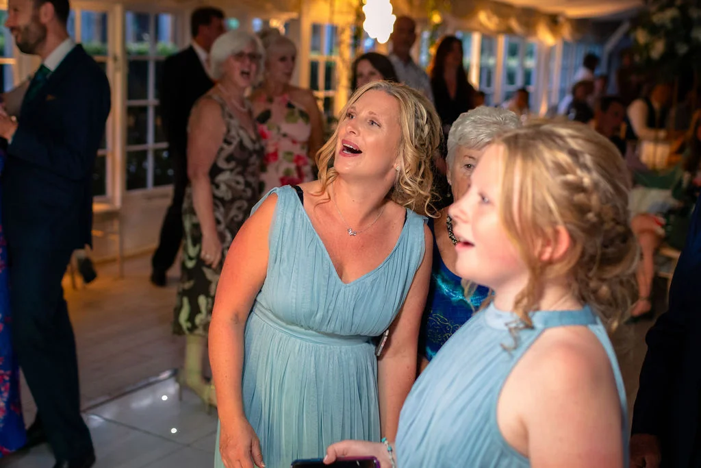 Wedding guests in blue dresses laughing happily during the speeches at a lively indoor evening reception.