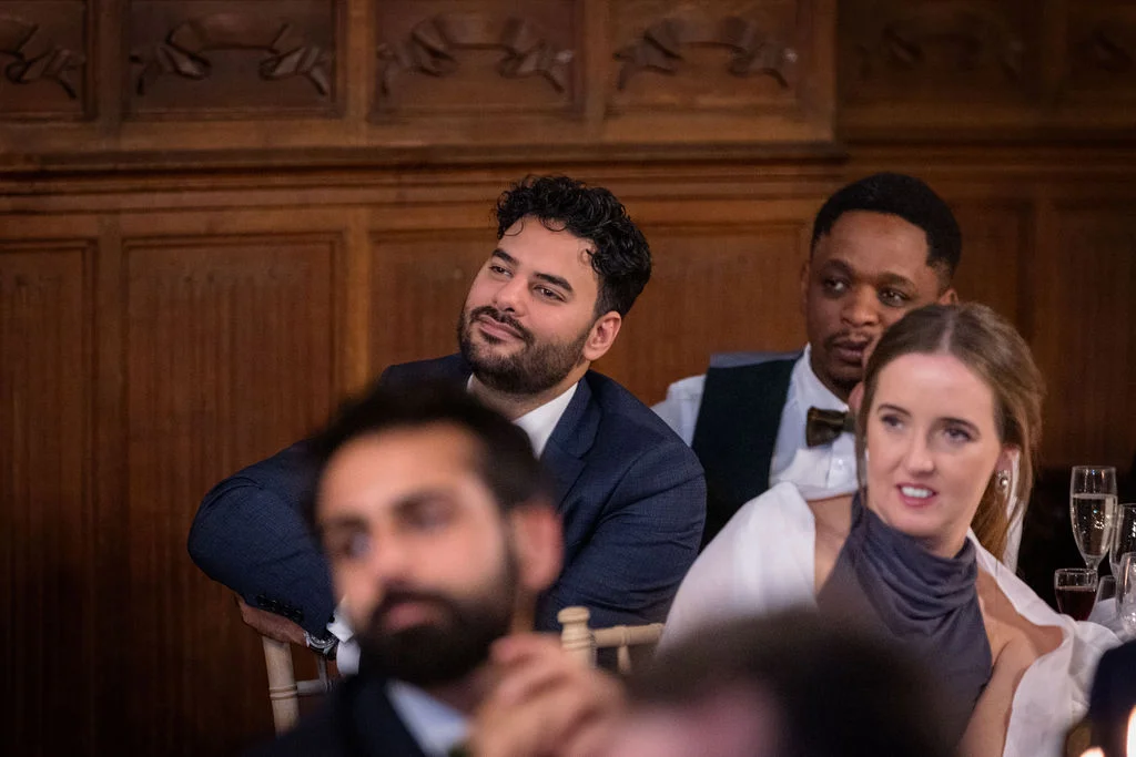 Wedding guests attentively watching speeches in a warm wood-panelled room with drinks on the tables.