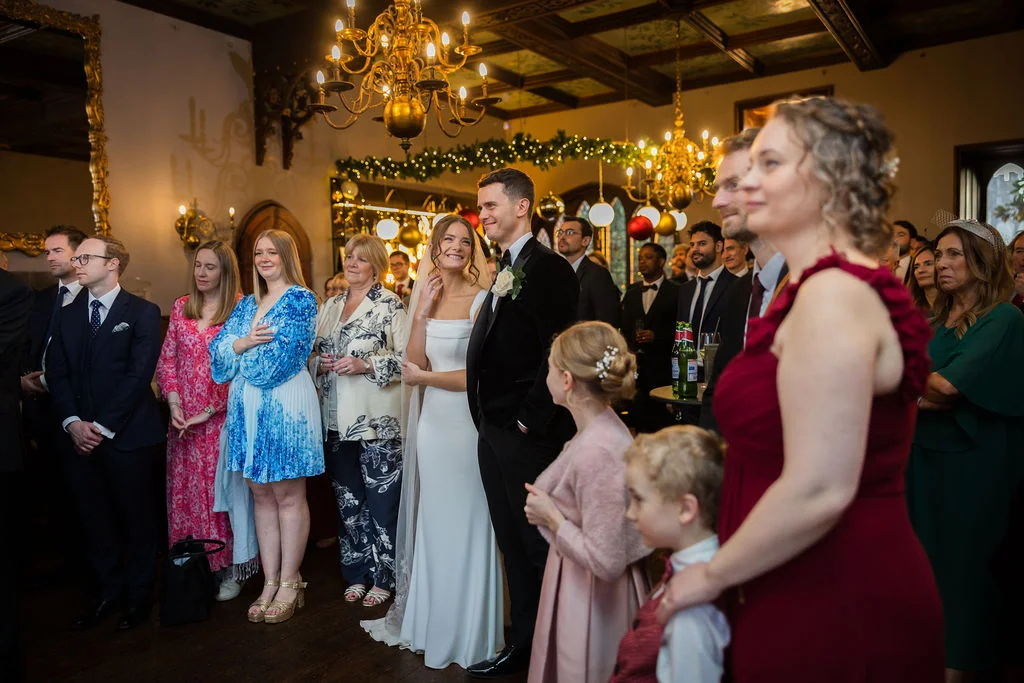 A newlywed couple stands together among a crowd of well-dressed guests inside a richly panelled hotel ballroom illuminated by grand chandeliers and fairy lights.