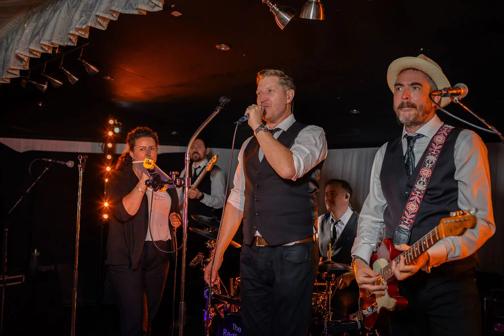 A lively wedding band performs on stage with vibrant lighting, featuring a lead singer with a microphone and a guitarist playing a red electric guitar.