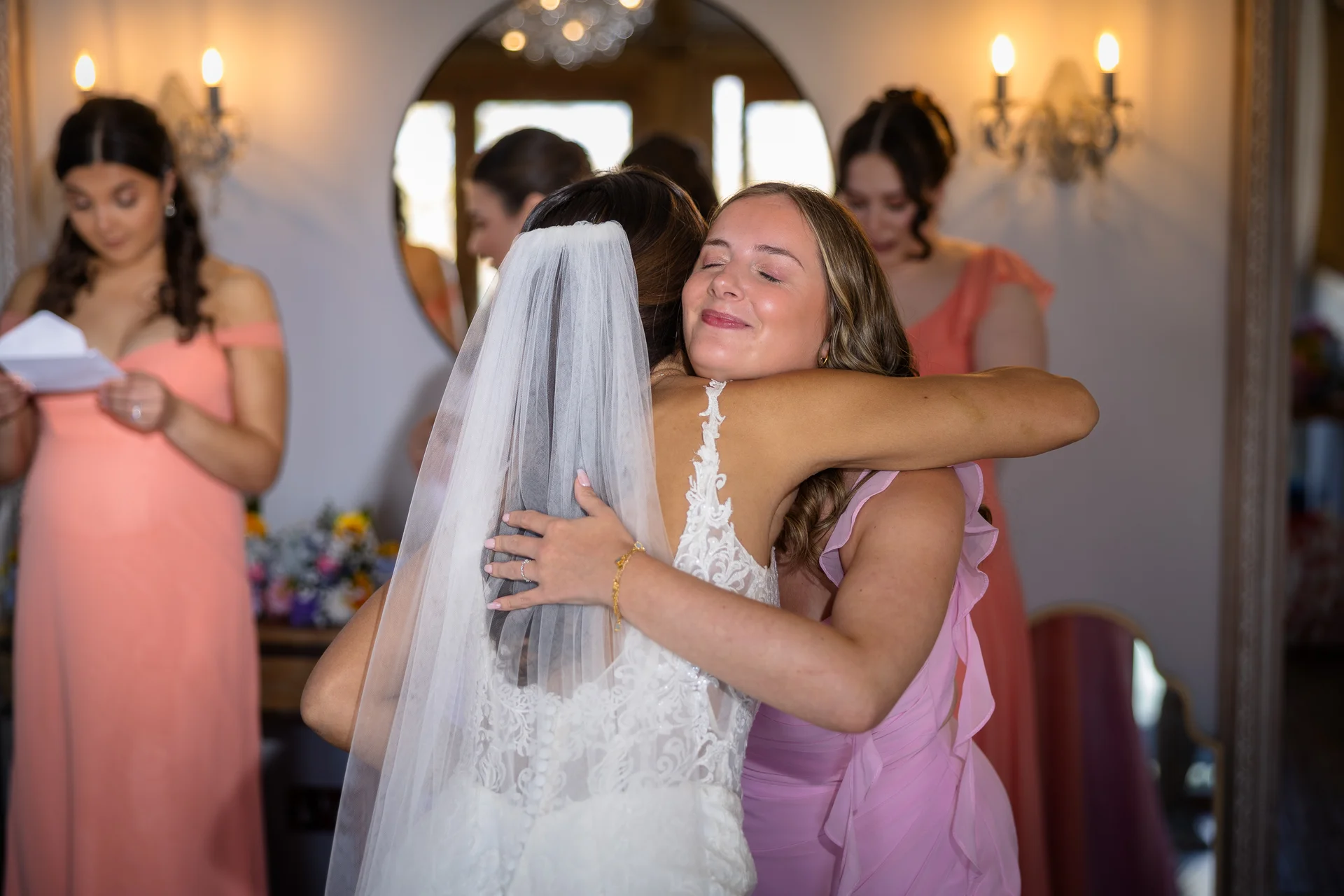 A joyful bride in a lace wedding dress embraces her bridesmaid during bridal preparations while other bridesmaids look on in the background.