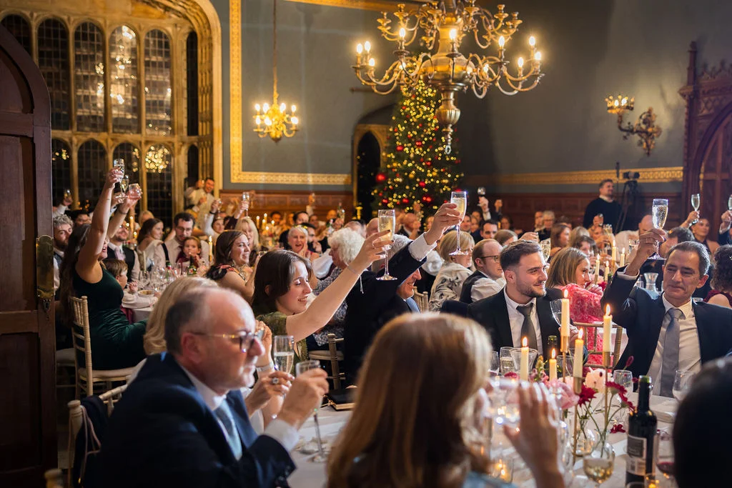 Wedding guests raise champagne glasses in a toast during a festive candlelit reception in a grand hall decorated with a Christmas tree.