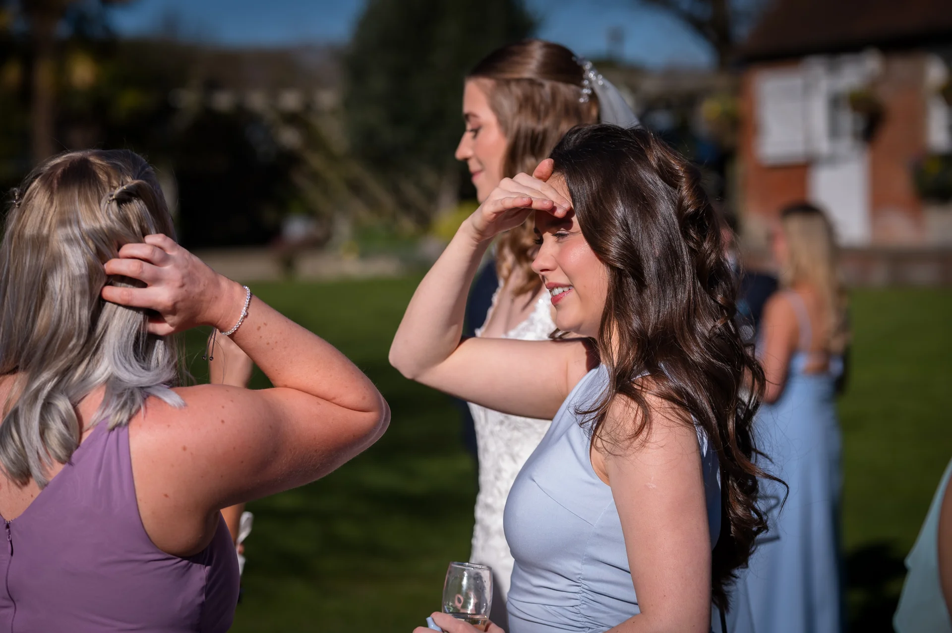 A smiling bridesmaid with dark wavy hair shades her eyes from the bright sunshine while chatting with the bride and friends outdoors in a manicured garden.