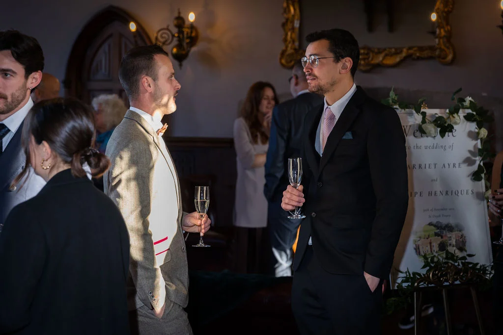 Wedding guests mingle and converse holding champagne flutes near a decorated welcome sign inside a historic venue.