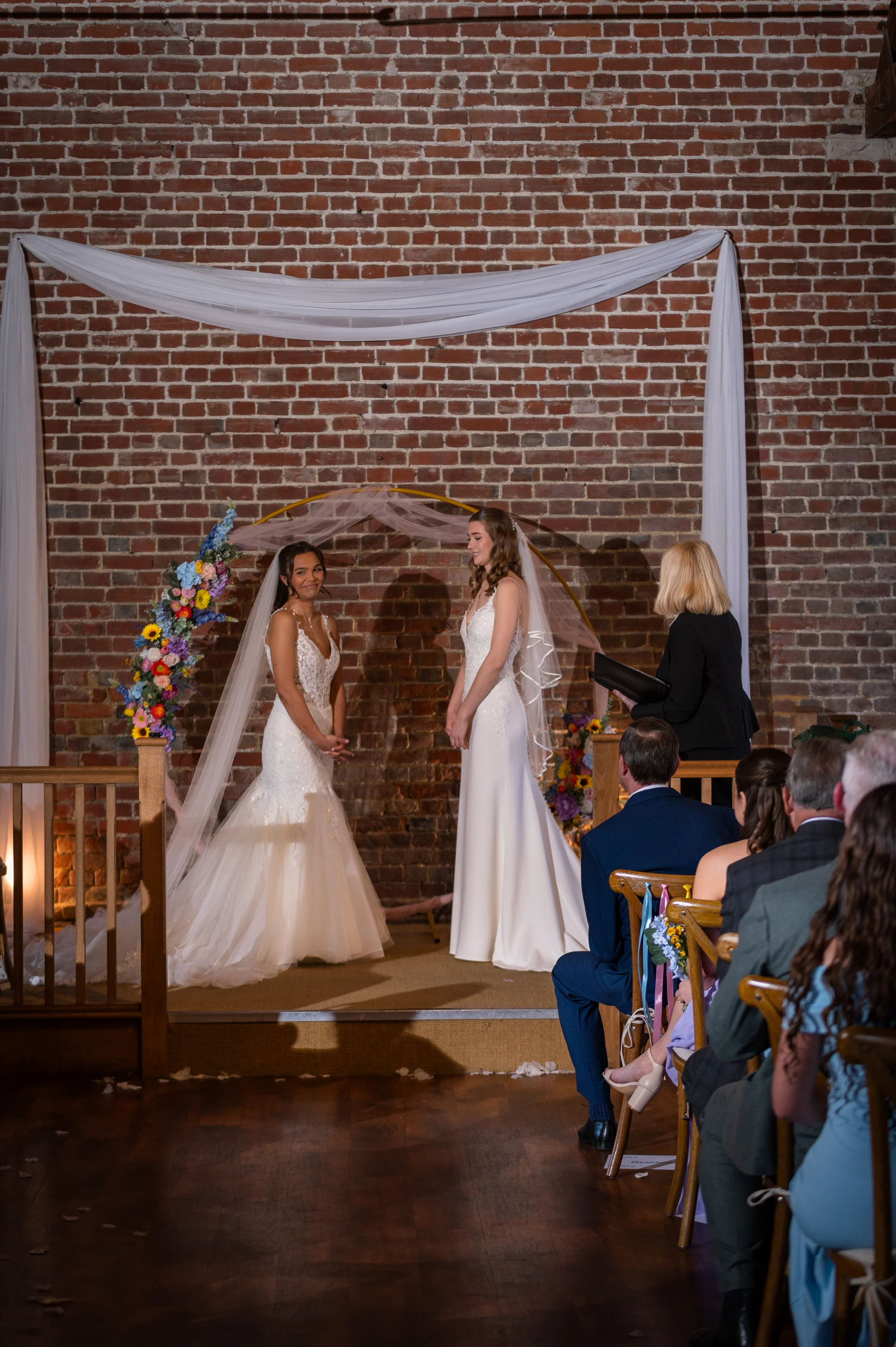 Two brides smile at each other during their wedding ceremony in an exposed brick barn venue, framed by a floral arch and white draping.