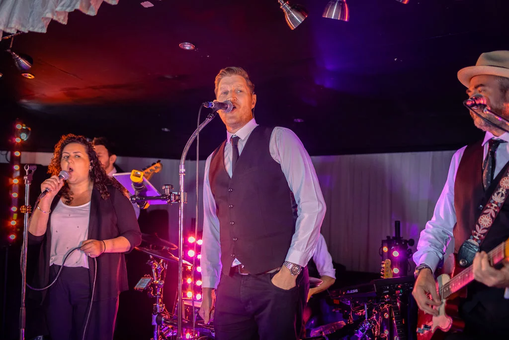 A live wedding band performs on stage under dramatic coloured lighting with a female vocalist and a male singer and guitarist.