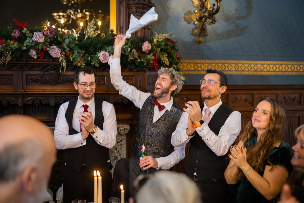 A group of enthusiastic guests and groomsmen cheer and wave a napkin enthusiastically during the wedding speeches inside a grand historic reception room.