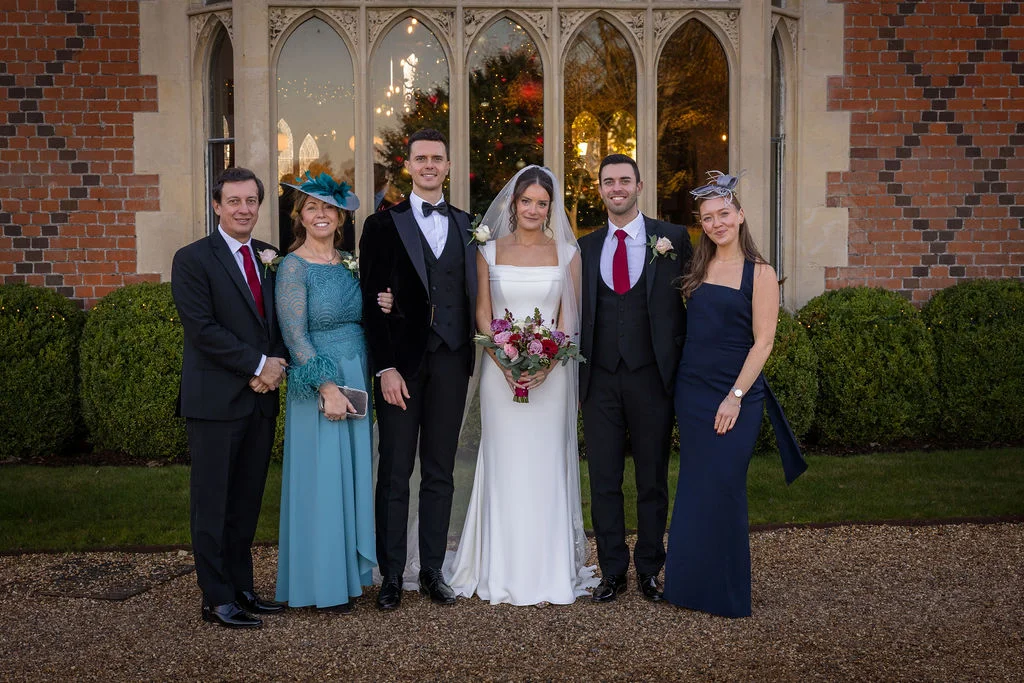 A bridal party and family members pose for a formal group portrait outside a stately home with tall arched windows at dusk.