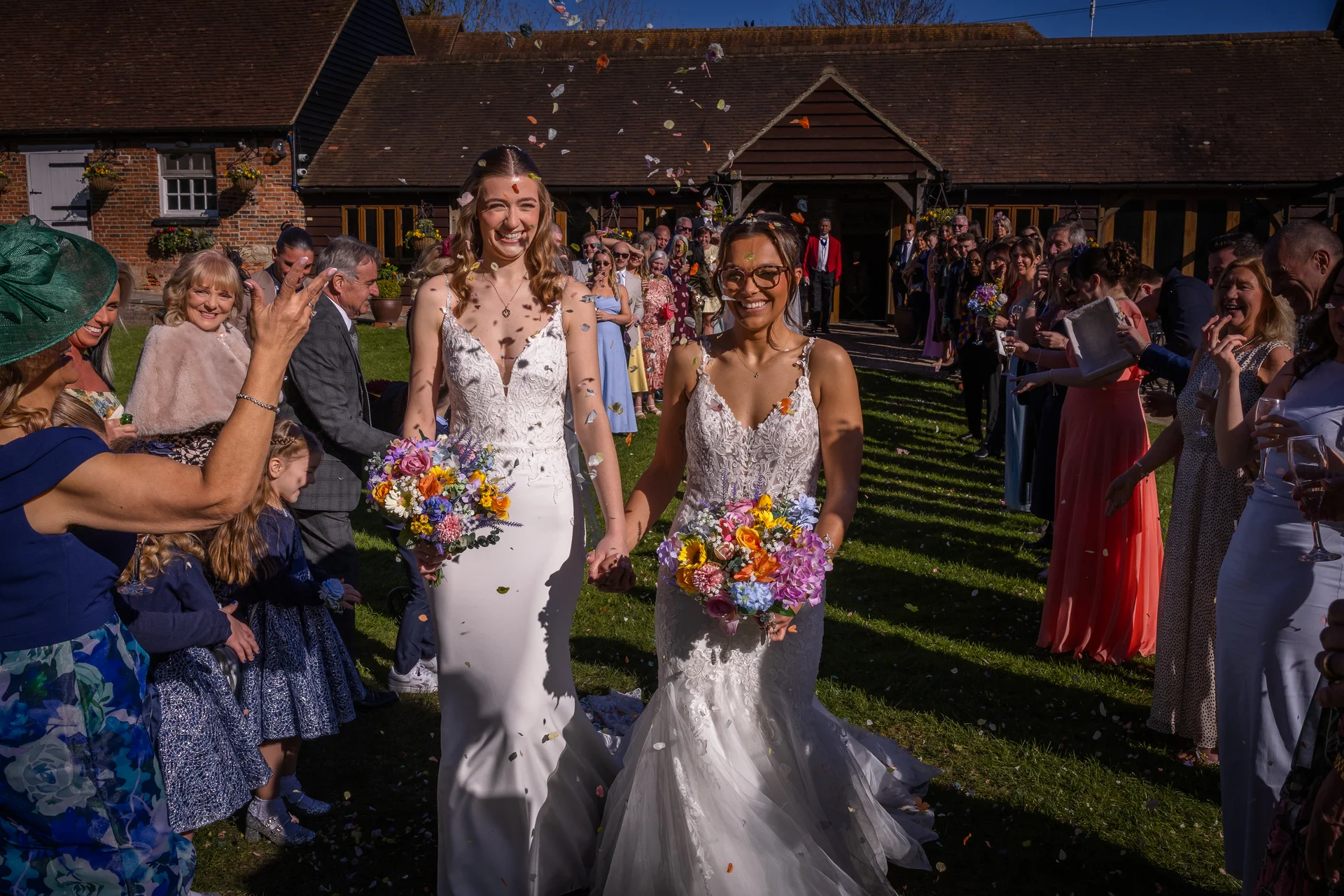 Two smiling brides holding colorful bouquets walk hand in hand through a joyful crowd throwing confetti outside a rustic barn venue on a sunny day.