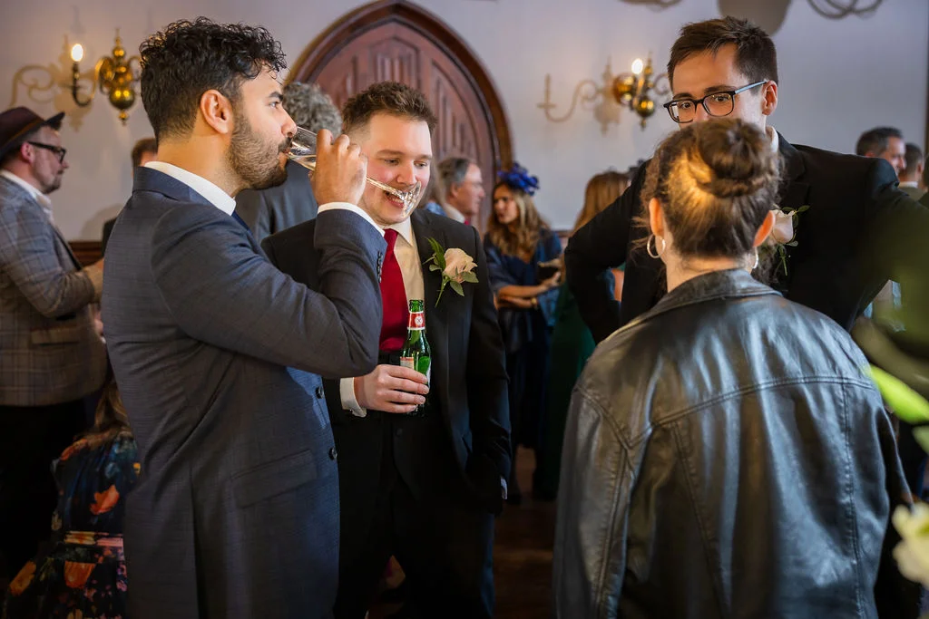 A group of wedding guests and wedding party members mingle indoors, laughing and raising drinks during the reception drinks.