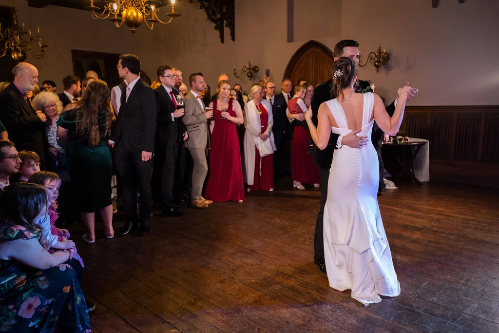 The newlywed couple share their first dance together on a wooden floor in a grand hall, surrounded by watching wedding guests.