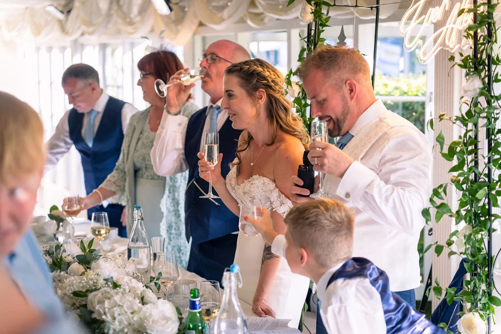 The newlywed couple and wedding party raise their champagne glasses for a toast at the top table decorated with lush white florals inside a draped marquee.