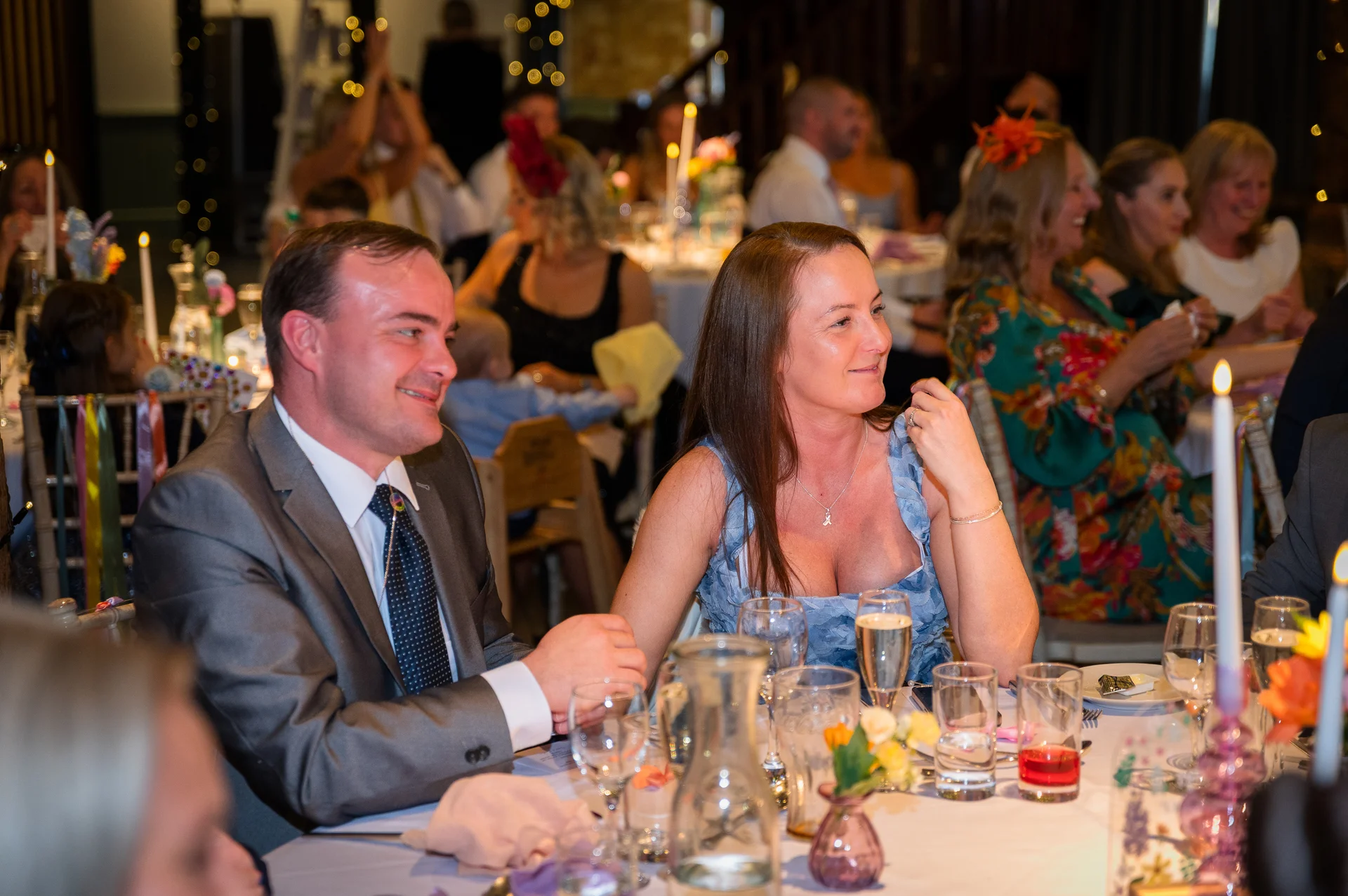 Wedding guests enjoy the wedding breakfast and speeches at a candlelit table, with a man in a grey suit and a woman in a blue dress smiling.