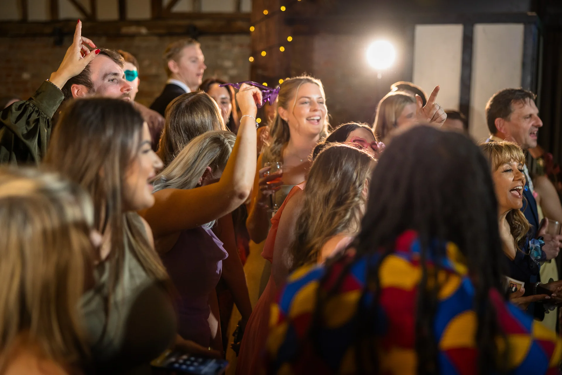 A lively crowd of wedding guests dances and cheers together on a dimly lit dance floor under string lights inside a rustic barn.