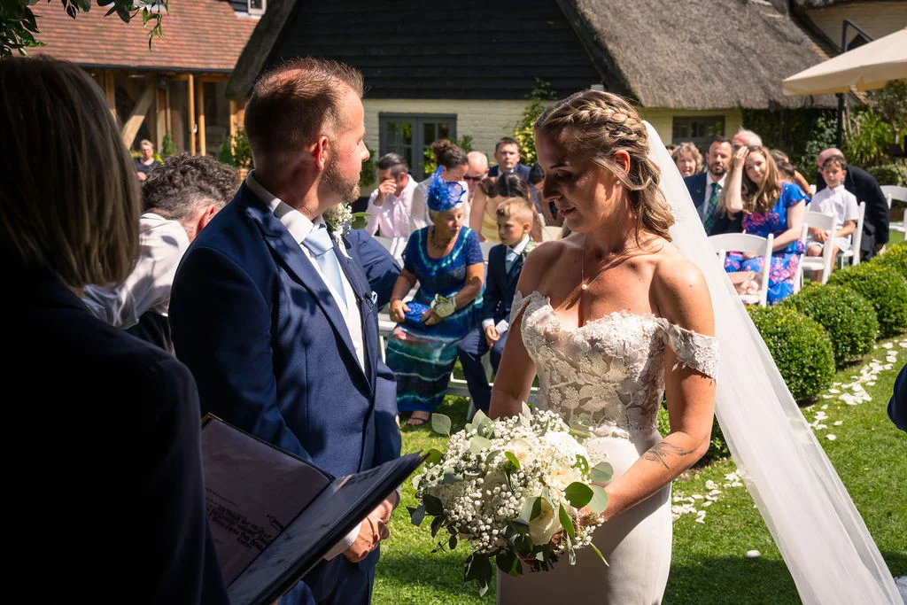A bride in an off-the-shoulder lace dress holding a white bouquet and a groom in a navy suit exchange vows during an outdoor summer ceremony on a sunny lawn.