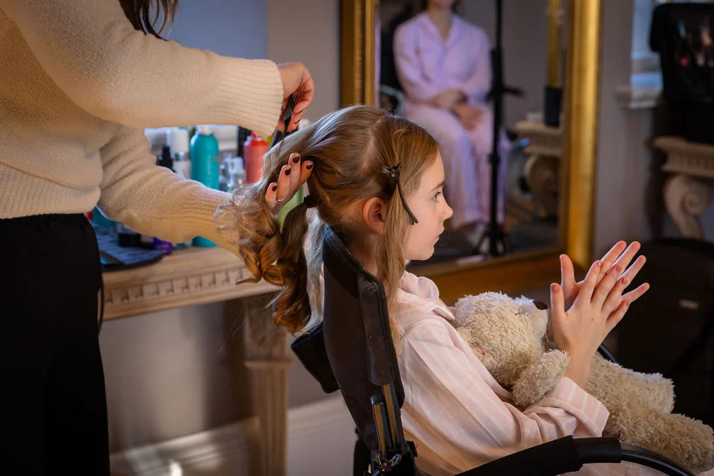 A young flower girl in matching pyjamas sits in a styling chair holding a teddy bear while having her hair styled during wedding morning preparations.