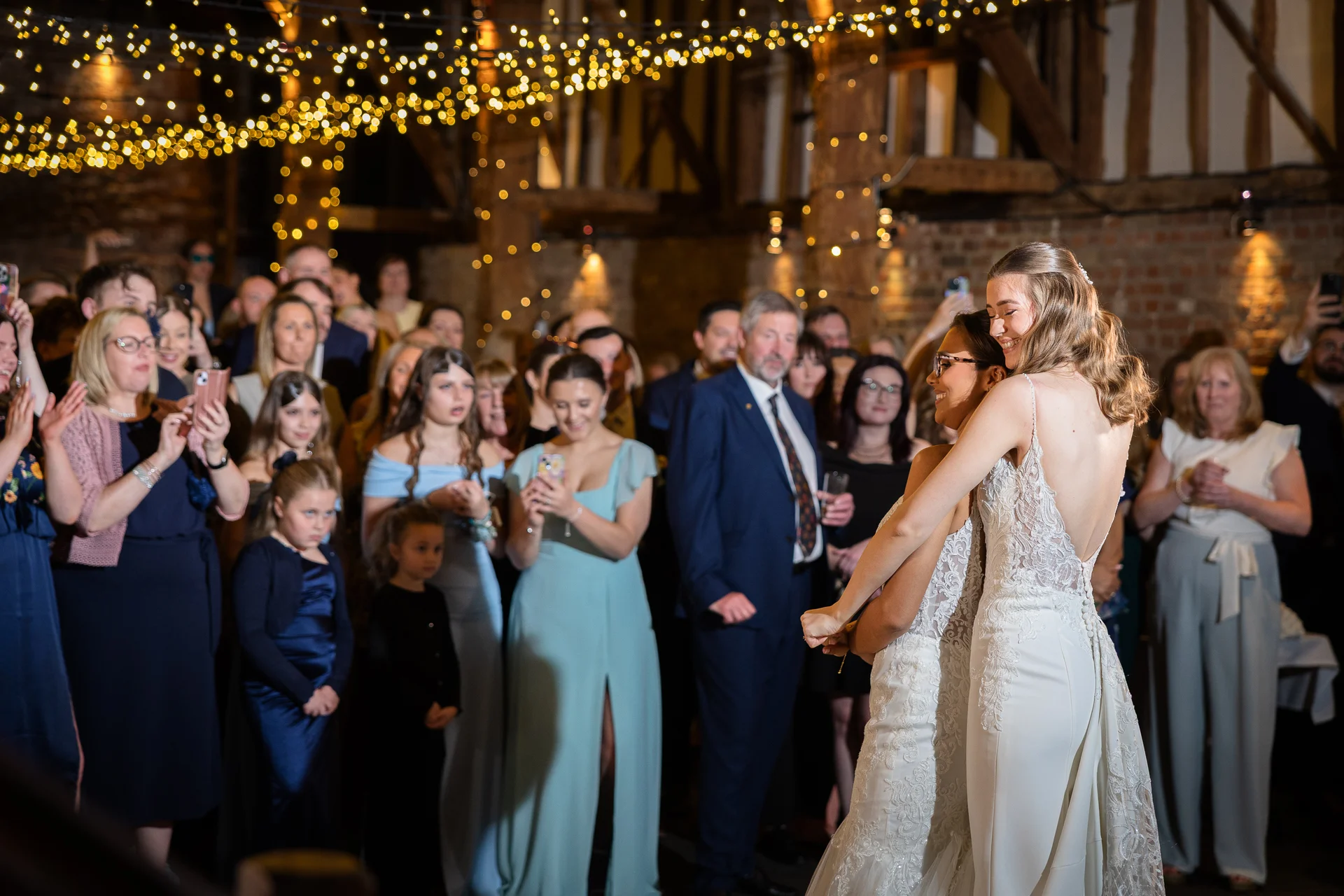 Two smiling brides embrace each other on the dance floor of a rustic oak-framed barn illuminated by a glowing canopy of fairy lights as guests watch happily.