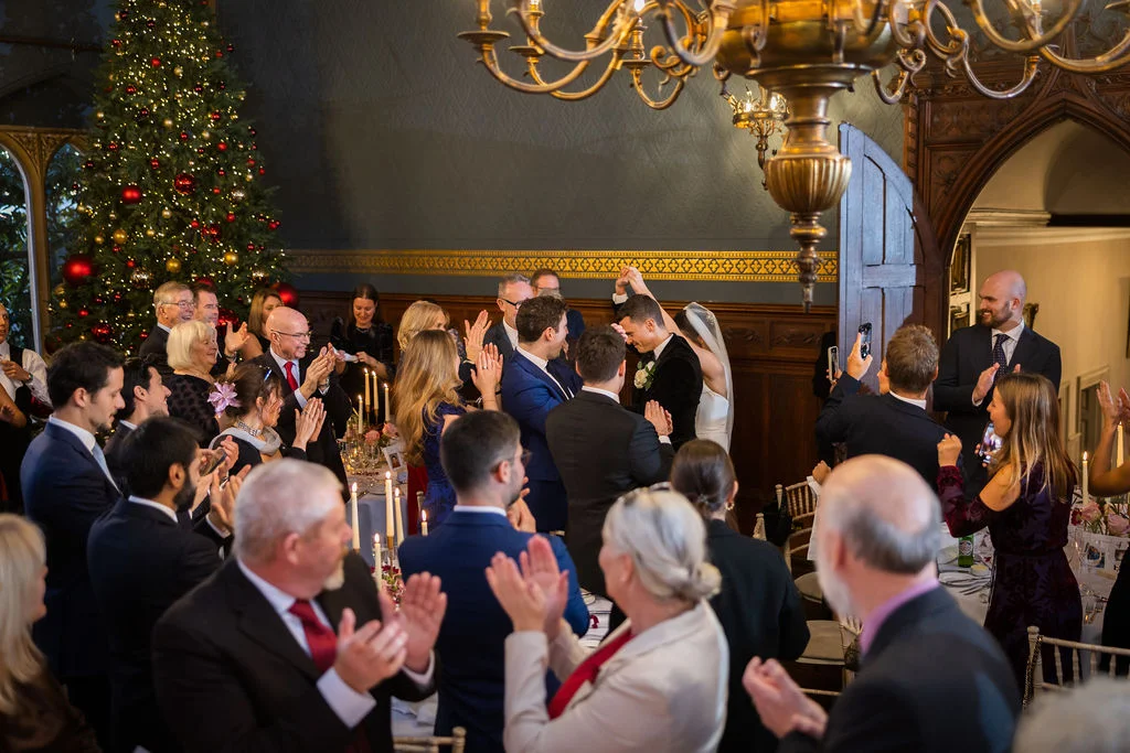 Newlyweds enter a grand country house reception room decorated with a festive Christmas tree, while seated guests applaud and stand to welcome them.