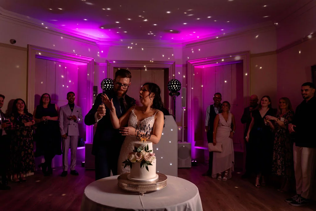A newlywed couple laugh together as they cut a white two-tier wedding cake decorated with fresh flowers, surrounded by guests in a reception room illuminated by vibrant purple lighting and disco ball effects.
