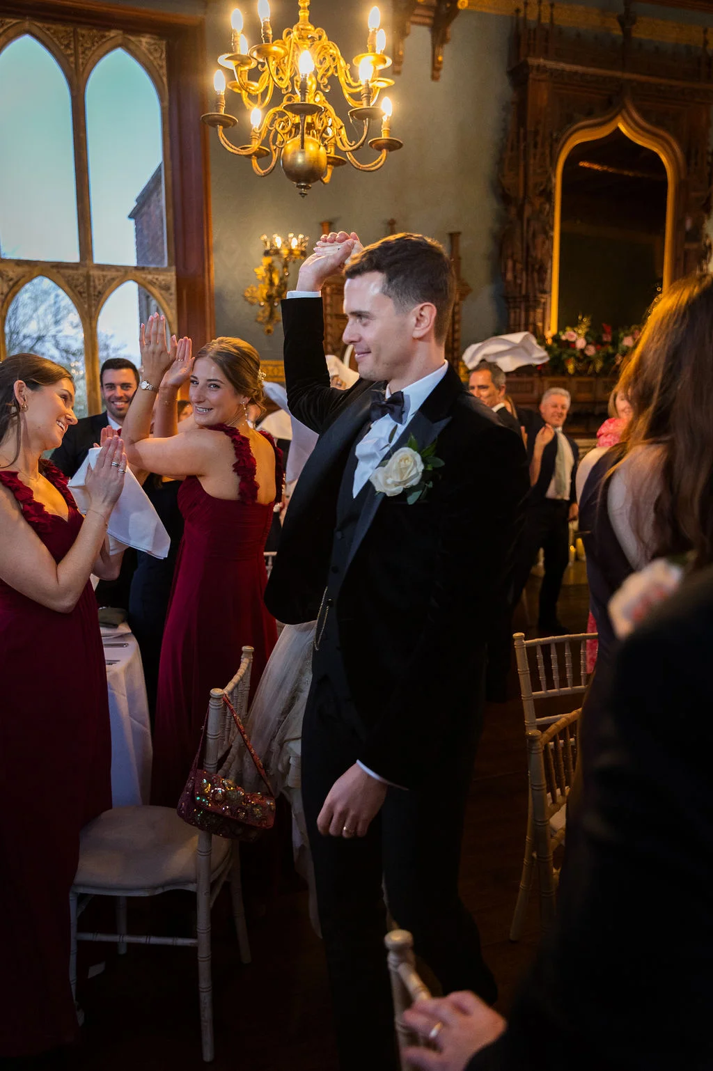 A groom in a black velvet tuxedo walks into a grand reception hall while cheering guests in burgundy dresses applaud him.