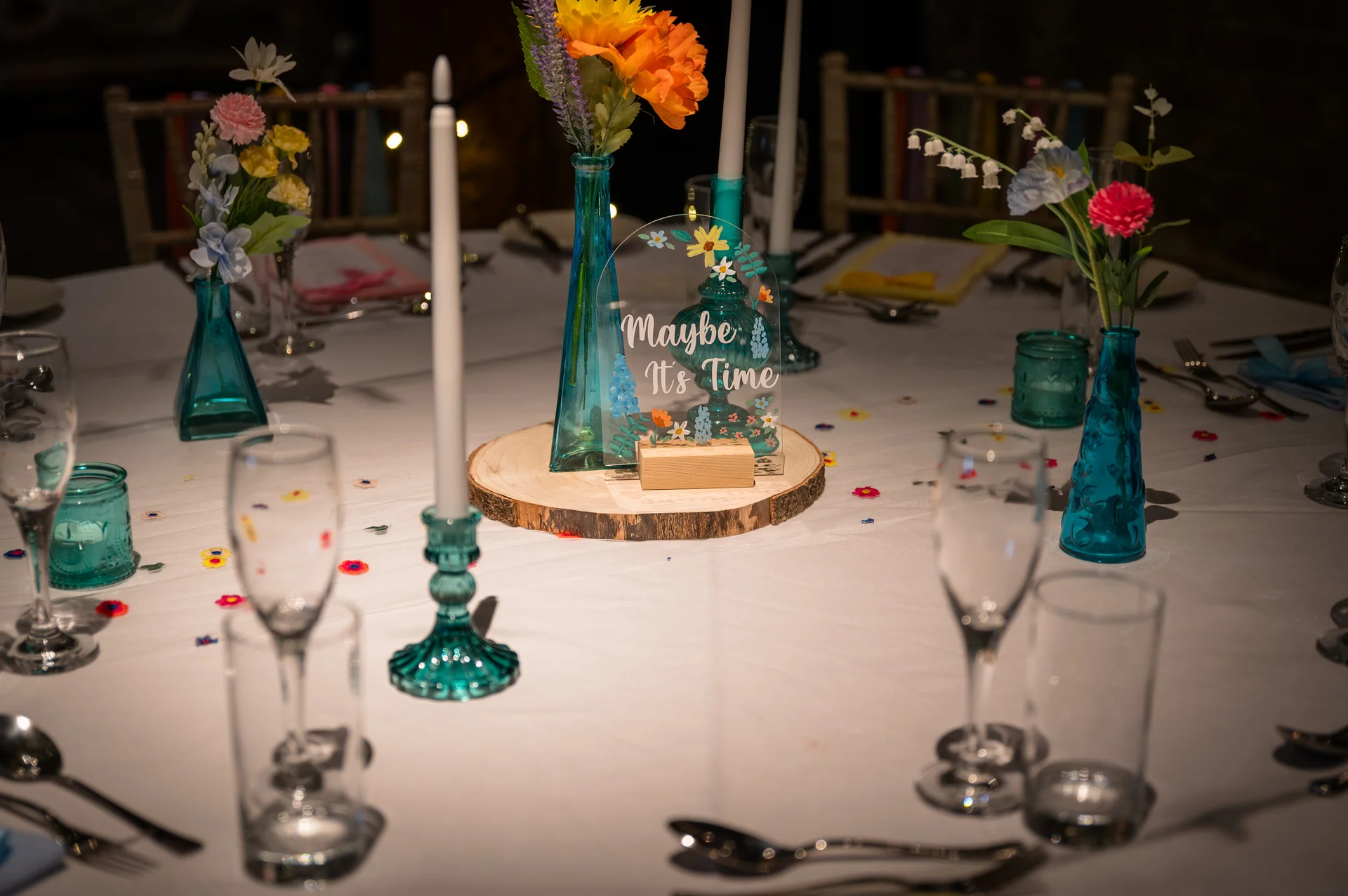 An elegantly set wedding round table features teal glassware, taper candles in matching holders, and bright floral bud vases surrounding a wooden slice centrepiece with an acrylic sign.