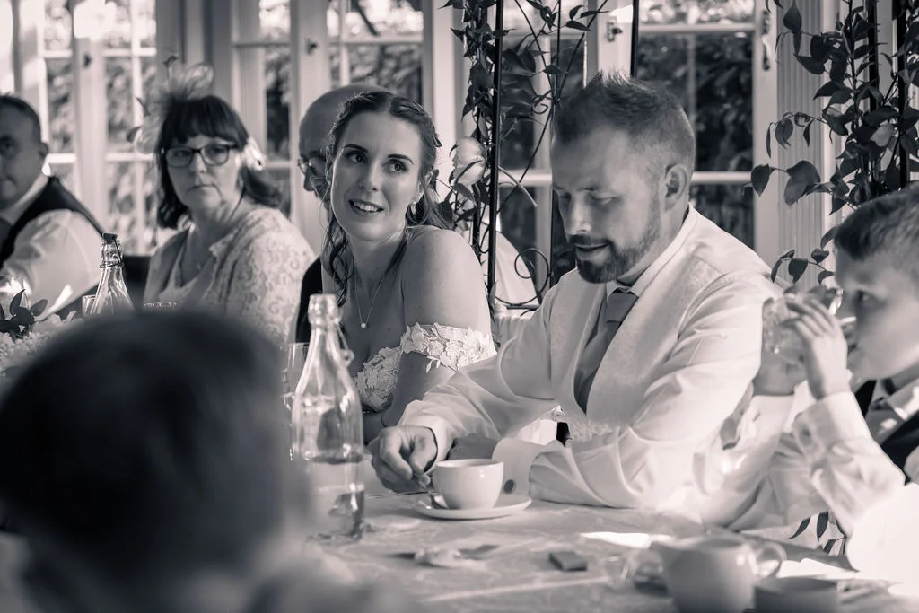 A candid black and white shot of the bride smiling warmly towards the groom during the wedding breakfast while guests sit at the table.