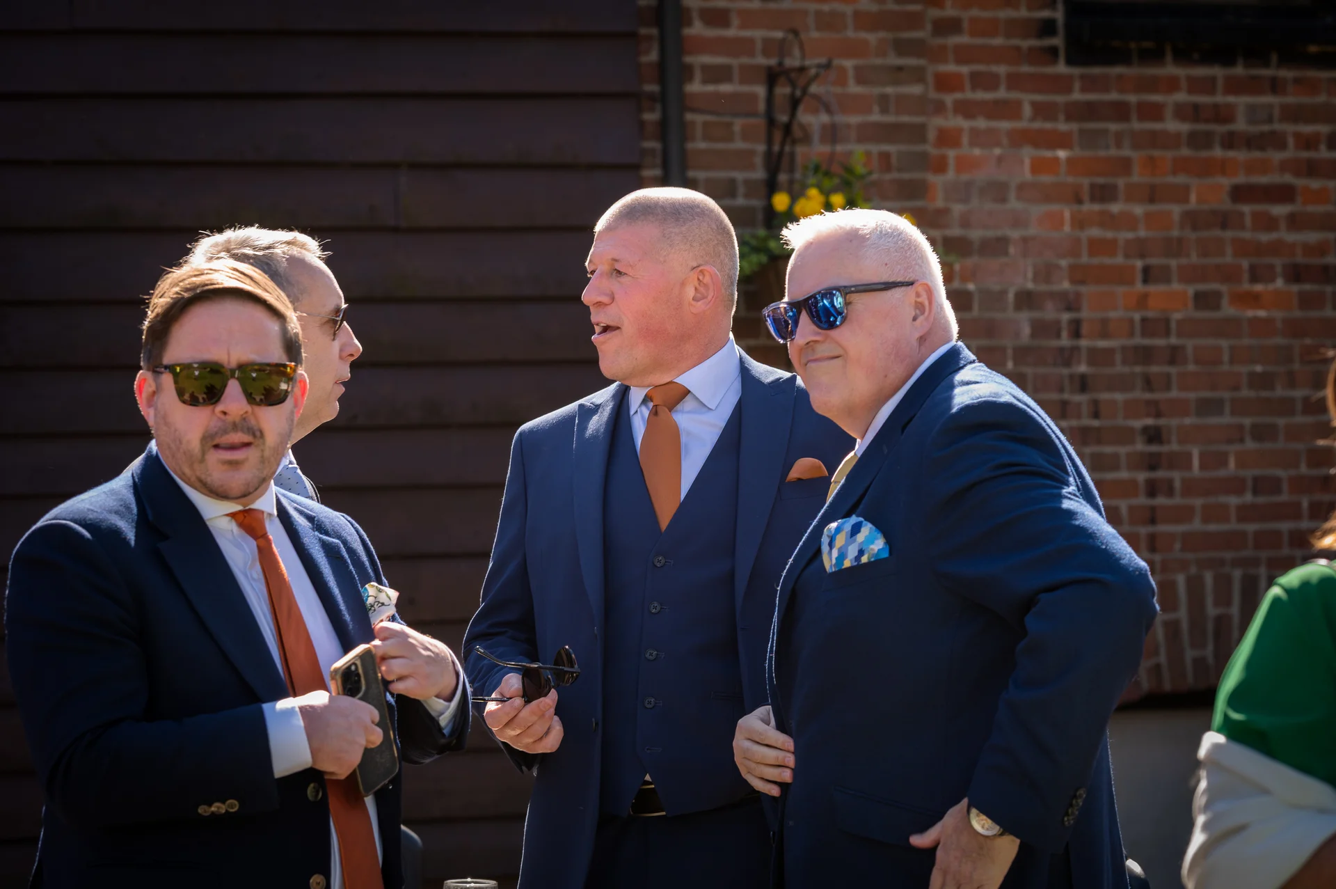 A group of male guests in tailored blue suits and orange ties mingle outside in the bright sunshine near an exposed brick wall.