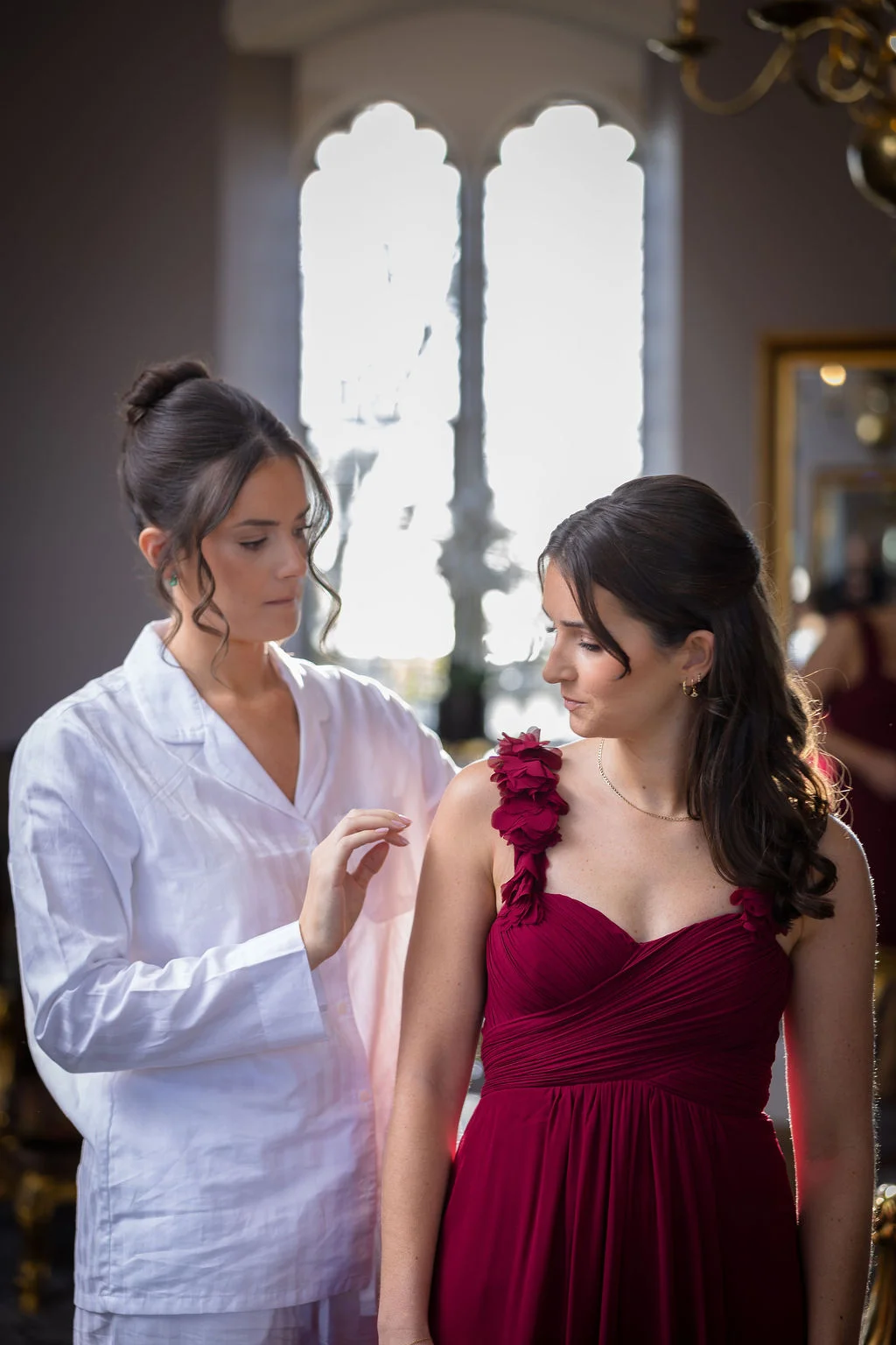 A bridesmaid in a matching white robe adjusts the strap of a stunning burgundy dress worn by another bridesmaid during morning preparations.