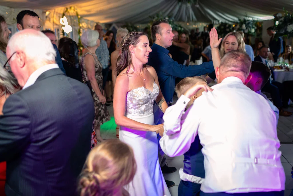 A joyous bride smiles while dancing with guests on a crowded marquee dance floor during an evening wedding reception.